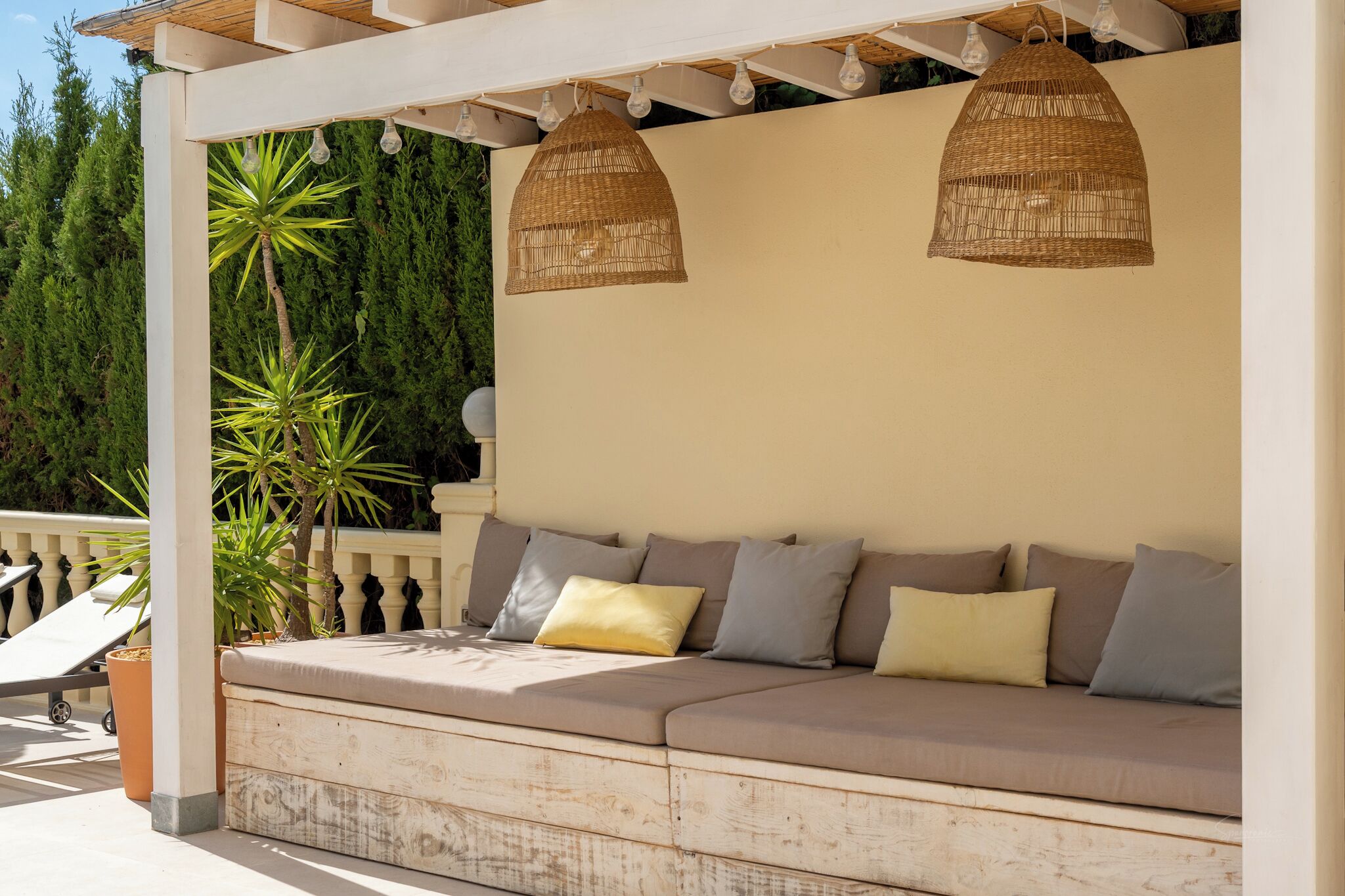 A covered outdoor seating area with a cushioned bench, wicker pendant lights, and potted plants.