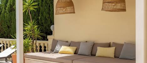 A covered outdoor seating area with a cushioned bench, wicker pendant lights, and potted plants.