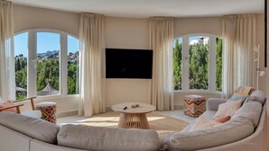 A living room with a large TV, a sofa, a round coffee table, and a view of greenery and buildings through large windows. - Large Villa with sea view in delightful Benissa Montemar (Costa Blanca North) (Benissa)