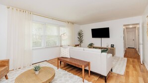 A modern living room with a white sofa, a wooden bench, a round table with a plant, and a flat-screen TV mounted on the wall. - Private Pool Oasis | 4BR Near Downtown Perry (Perry)