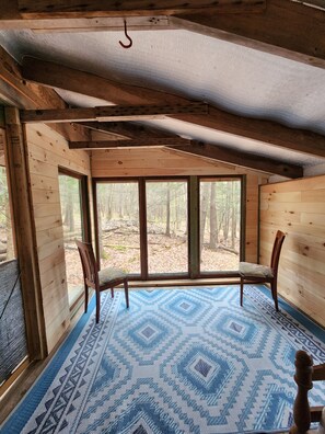 A cabin interior with a wooden floor, a blue patterned rug, and a large window overlooking a forest. - Writer's Den: Rustic Retreat Camping Cabin (Freeville)