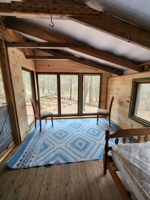 A cabin with a large window, a bed, and a blue patterned rug. - Writer's Den: Rustic Retreat Camping Cabin (Freeville)