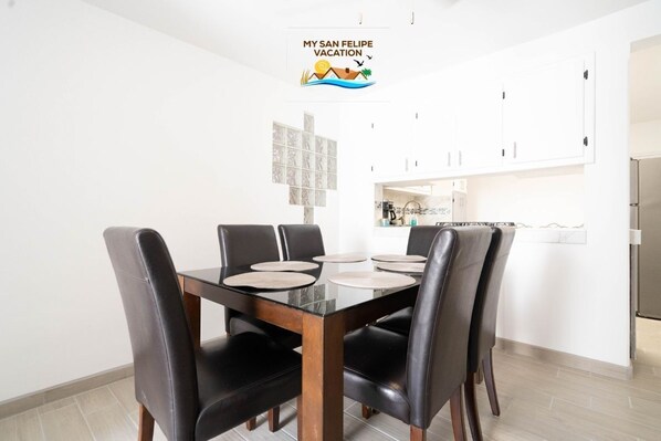 Un coin repas avec une table en verre et des chaises noires, une cuisine en arrière-plan et une pancarte sur laquelle on peut lire : « MES VACANCES À SAN FELIPE ».