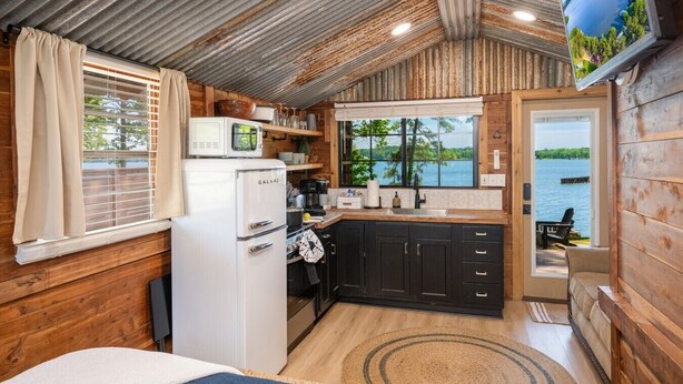 A compact kitchen with a refrigerator, microwave, and stove, wooden walls, and a view of a lake.