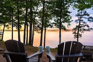 Een terras aan het meer met Adirondack-stoelen en een tafel, met uitzicht op een kalm meer en een bebost gebied.