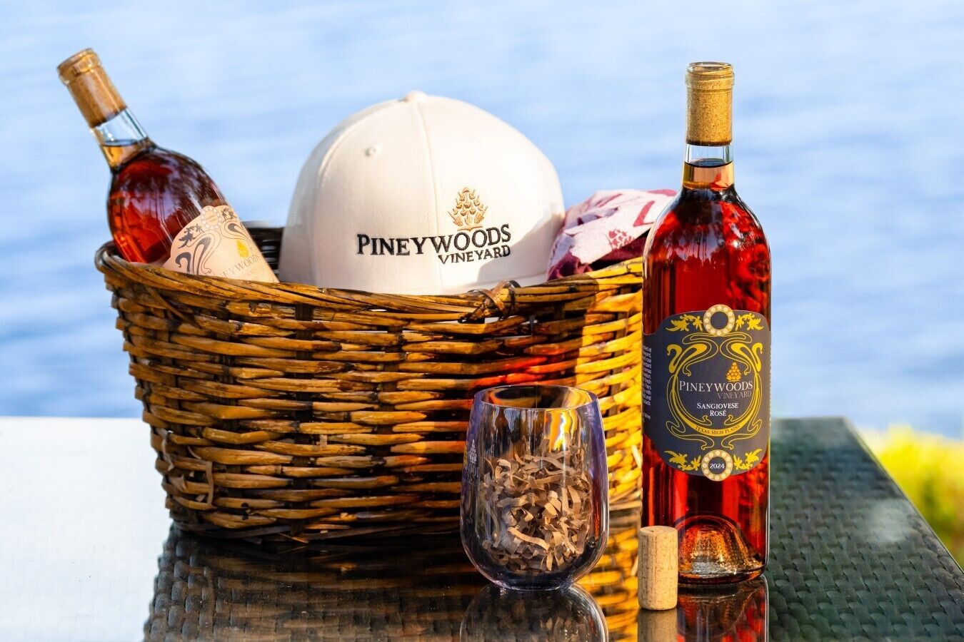A wicker basket containing a white cap, two bottles of Piney Woods Vineyards Sangiovese, and a glass with wood shavings.