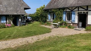 Il s’agit d’un bâtiment de style traditionnel, semblable à un chalet, avec un toit de chaume, une allée gravillonnée et un petit jardin agrémenté d’une table et de chaises.