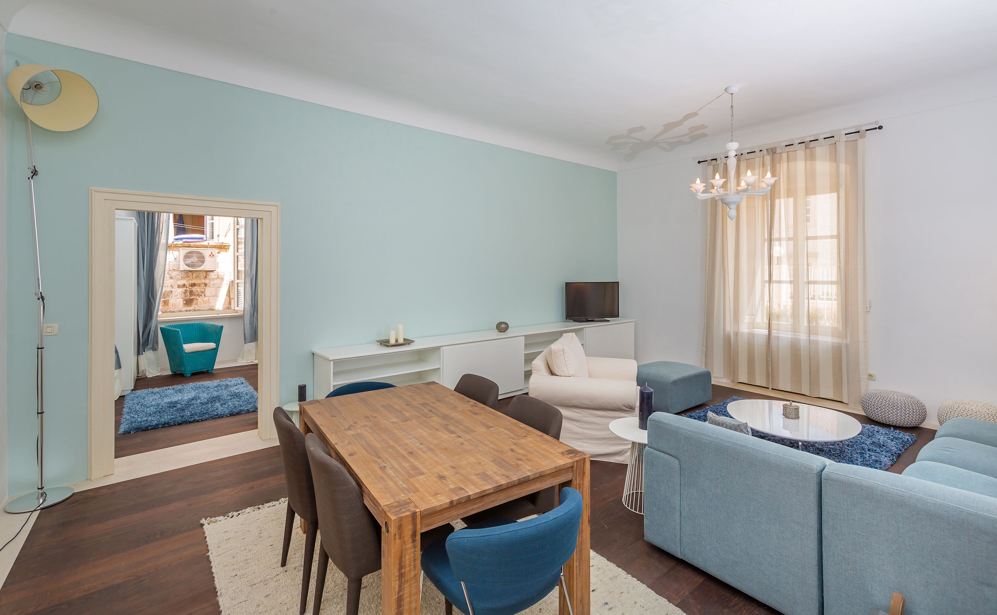 Un salon moderne avec une table à manger en bois, des chaises bleues, un canapé bleu clair et un téléviseur posé sur un meuble blanc.