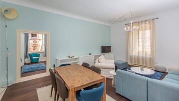 A modern living room with a wooden dining table, blue chairs, a light blue sofa, and a TV on a white stand.