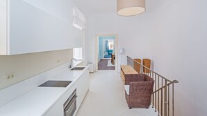 Une cuisine moderne avec des meubles blancs, un four encastré et un évier. On y trouve un banc en osier et une lampe près d’un escalier.