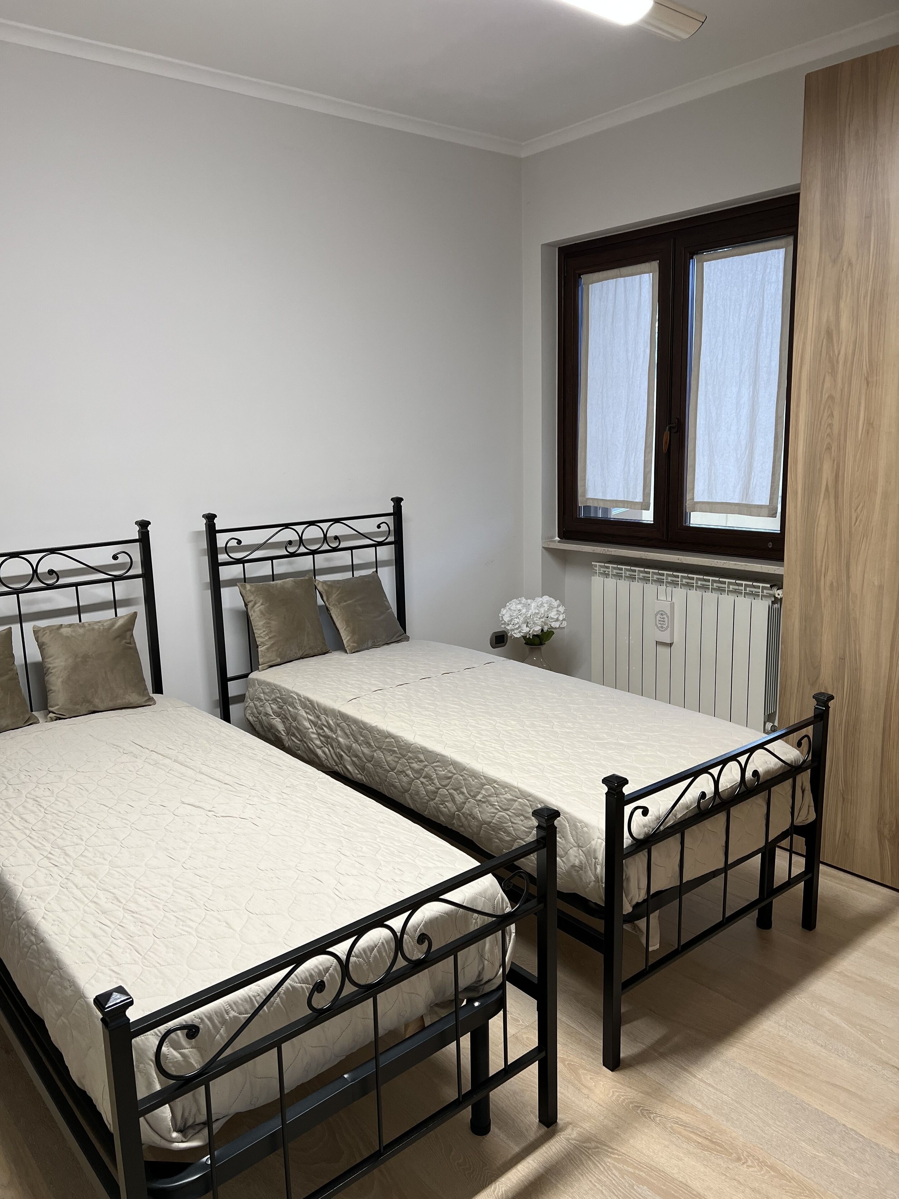 A bedroom with two single beds, a window with curtains, a radiator, and a wooden door.