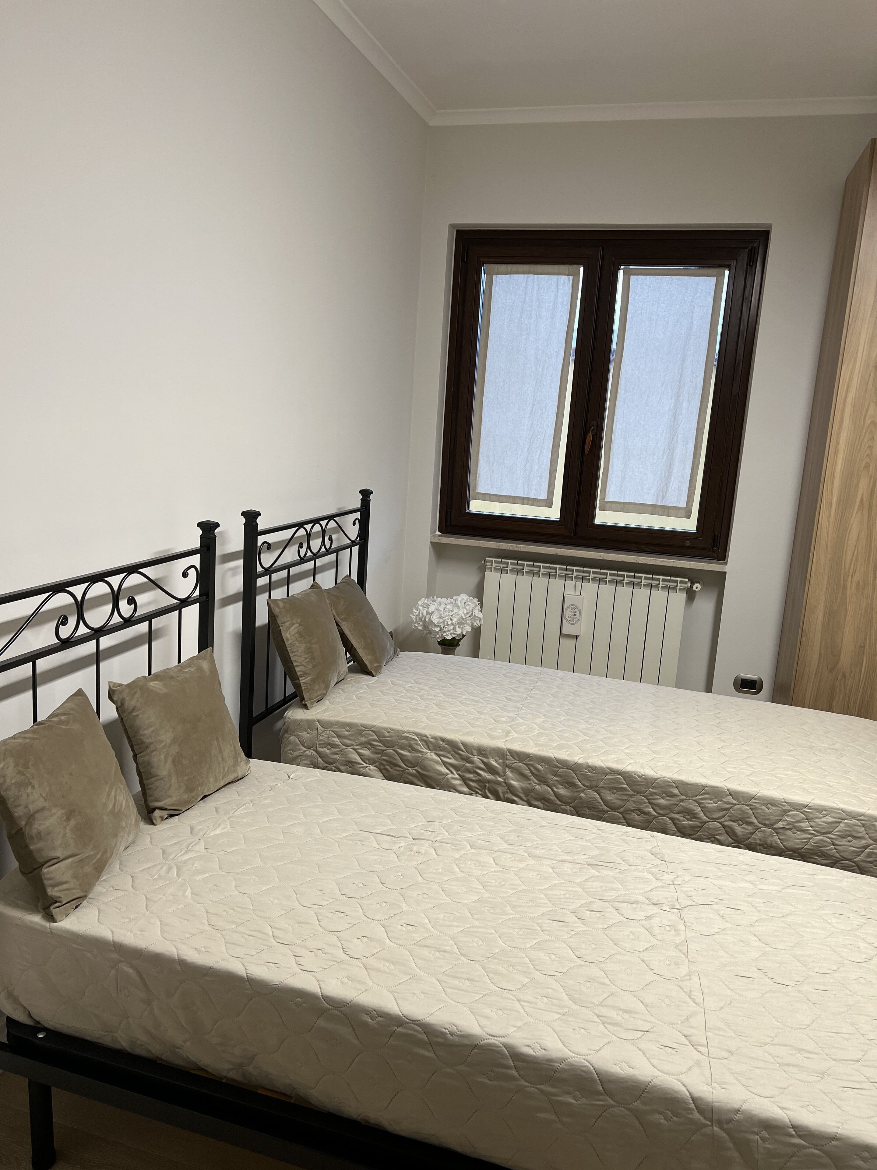 A bedroom with two beds, a window with blinds, a radiator, and a white flower vase.