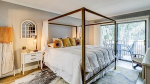 A four-poster bed with a canopy, a bedside table with a lamp, a window with a view of trees, and a balcony with chairs.
