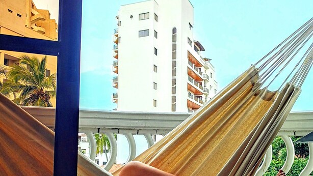 Une personne allongée dans un hamac sur un balcon dont le sol est carrelé, avec en arrière-plan la vue d’un bâtiment blanc.