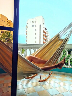 Une personne allongée dans un hamac sur un balcon dont le sol est carrelé, avec en arrière-plan la vue d’un bâtiment blanc.