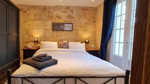 Ein ordentlich bezogenes Bett mit weißer Daunendecke, zwei Nachttischlampen, ein gerahmtes Kunstwerk an der Wand und ein großes Fenster mit Vorhängen.