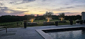 A pool area with stone flooring, a water feature, and three lit fire pits.