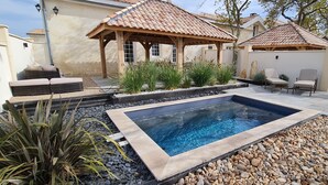 Ein Pool mit Holzterrasse, umgeben von Felsen und Pflanzen, sowie ein überdachter Sitzbereich mit Stühlen.