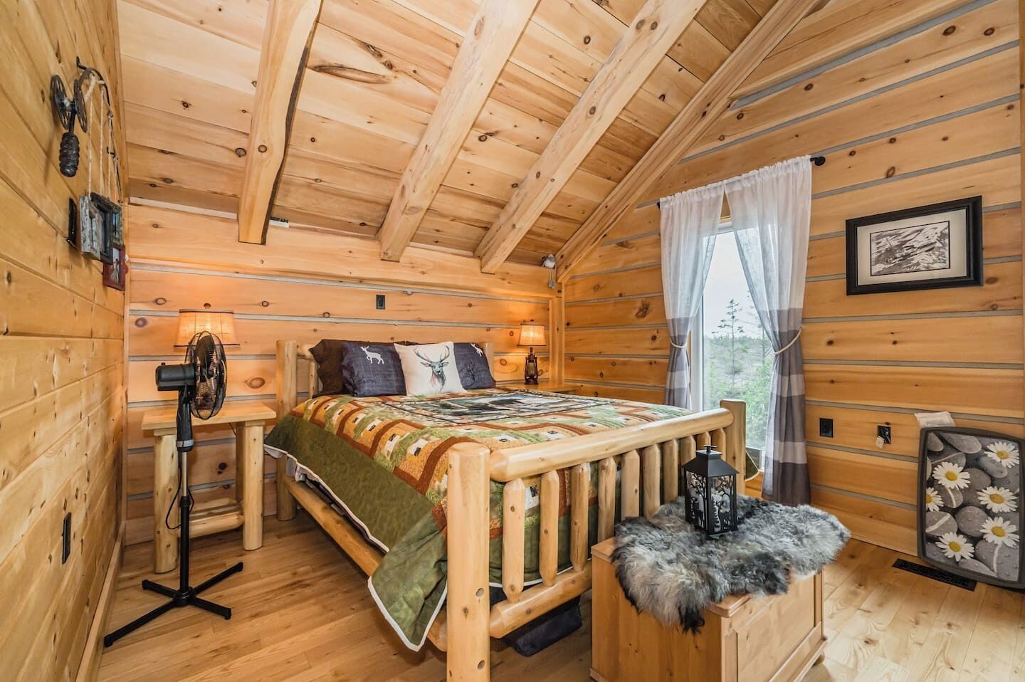 A cozy log cabin bedroom with a wooden bed, a standing fan, and a rustic decor including a lantern and a framed picture.