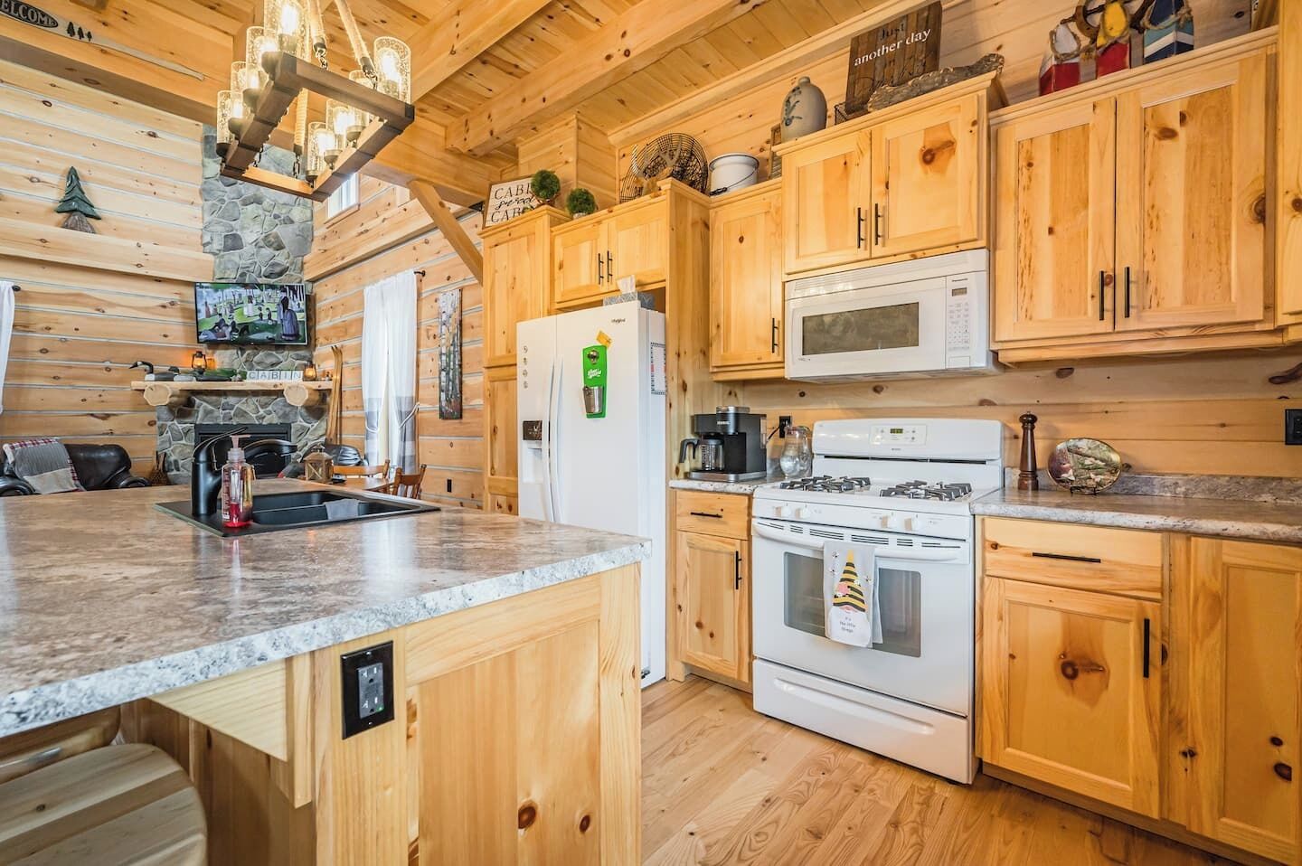 A wooden cabin-style kitchen with a stone fireplace, wooden cabinets, a granite countertop, a microwave, a stove, and a refrigerator.