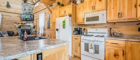 A wooden cabin-style kitchen with a stone fireplace, wooden cabinets, a granite countertop, a microwave, a stove, and a refrigerator.