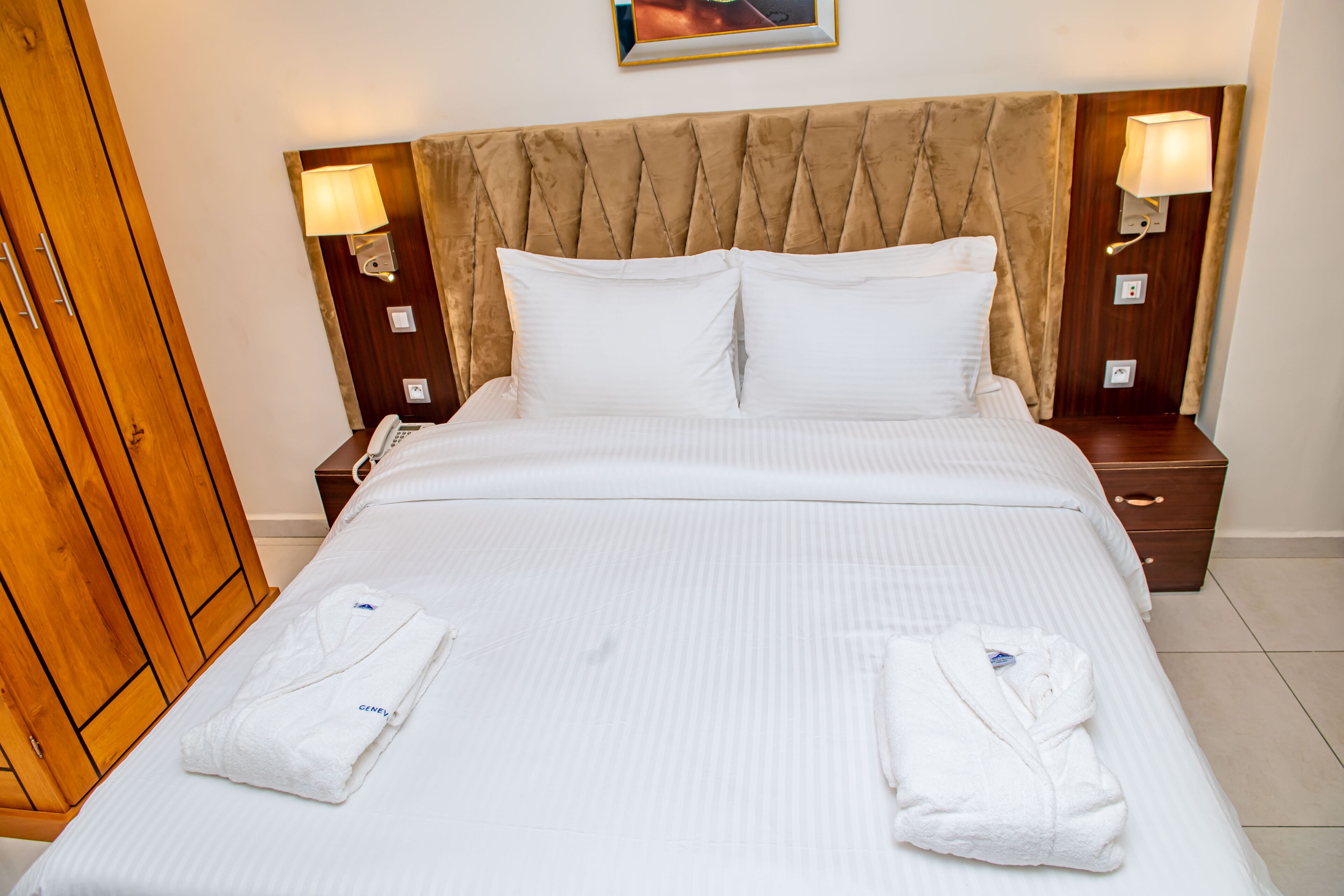 Una cama bien hecha con sábanas blancas y dos toallas dobladas encima. Hay dos mesitas de noche con lámparas, un armario de madera y un cuadro en la pared.