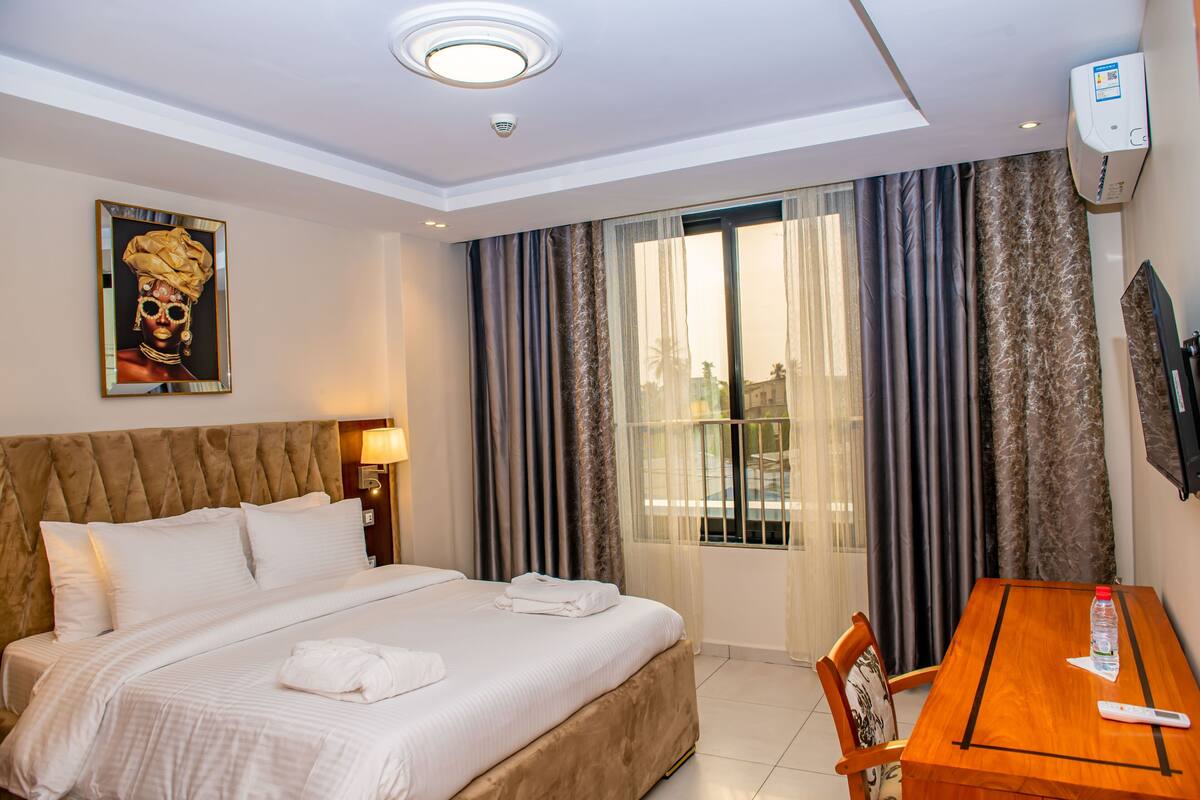 A hotel room with a bed, a table, a chair, a painting, and a window with curtains.
