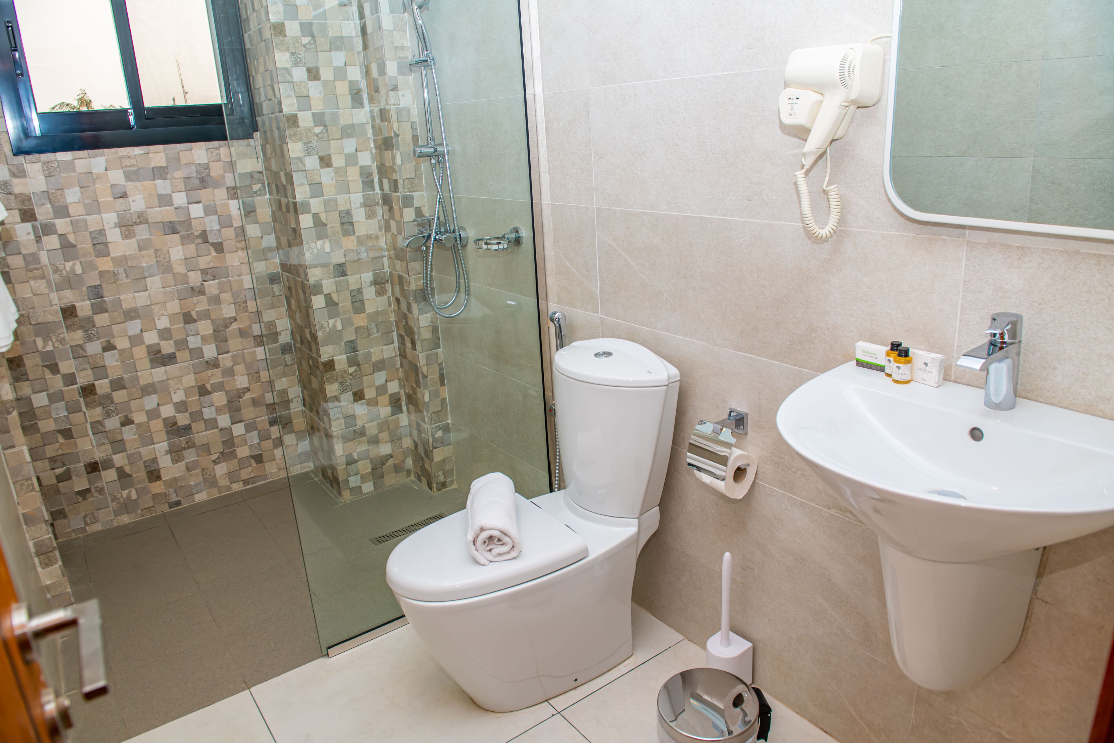 A bathroom with a glass shower enclosure, a toilet, a sink, and a hairdryer mounted on the wall.