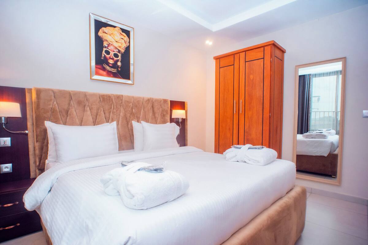 A hotel room with a large bed, a wooden wardrobe, a framed picture on the wall, and a mirror reflecting the bed.