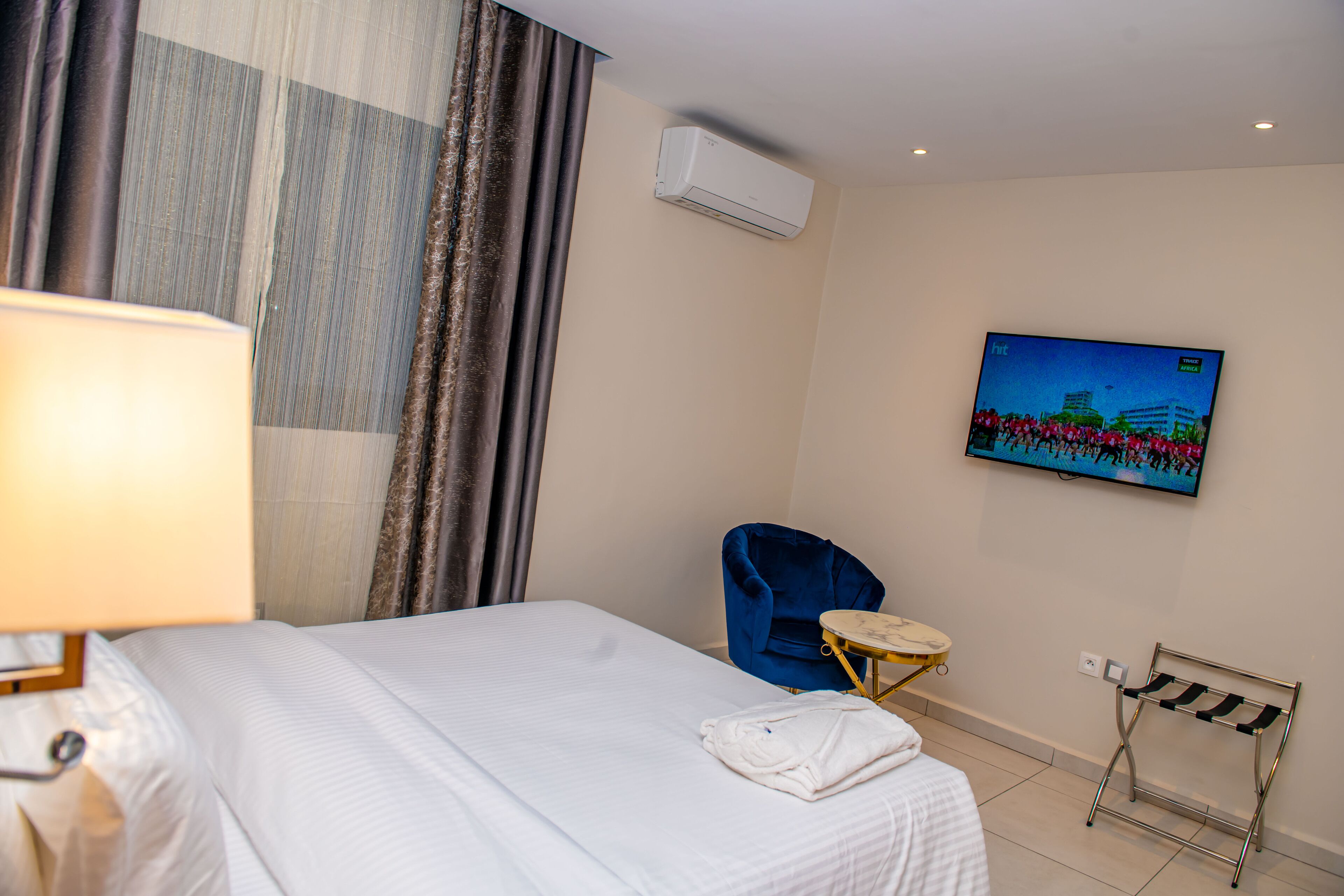 Une chambre d’hôtel avec un lit, une chaise bleue, un téléviseur fixé au mur et un appareil de climatisation.