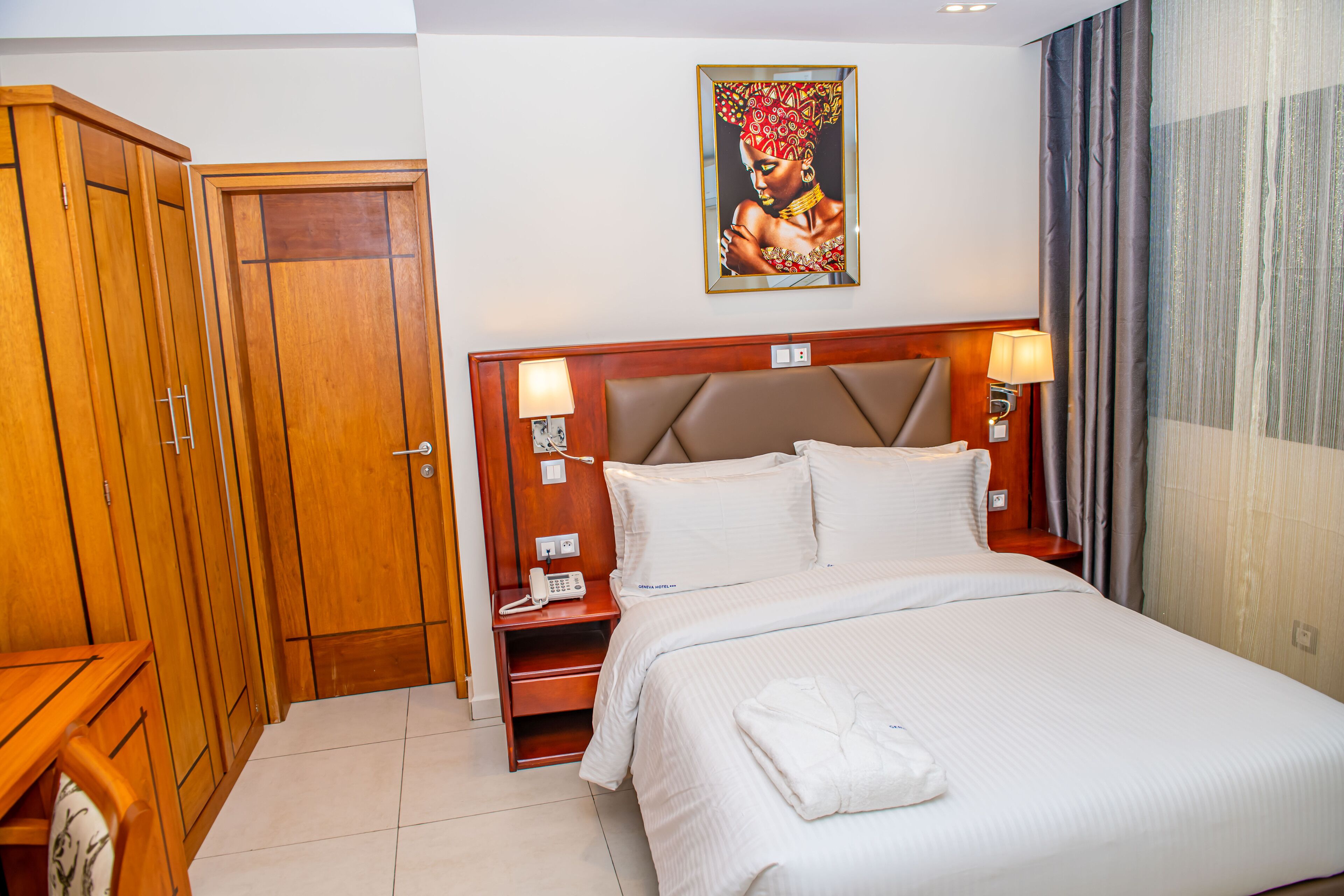 A hotel room with a large bed, wooden wardrobe, bedside lamps, and a painting on the wall.