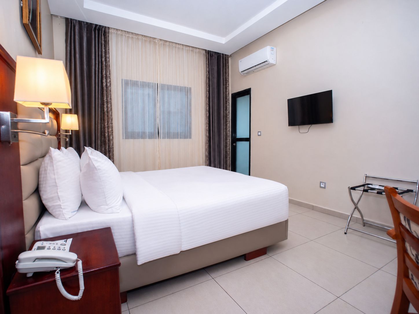 A hotel room with a bed, bedside lamp, a telephone, an air conditioning unit, a television, and a small desk with a chair.