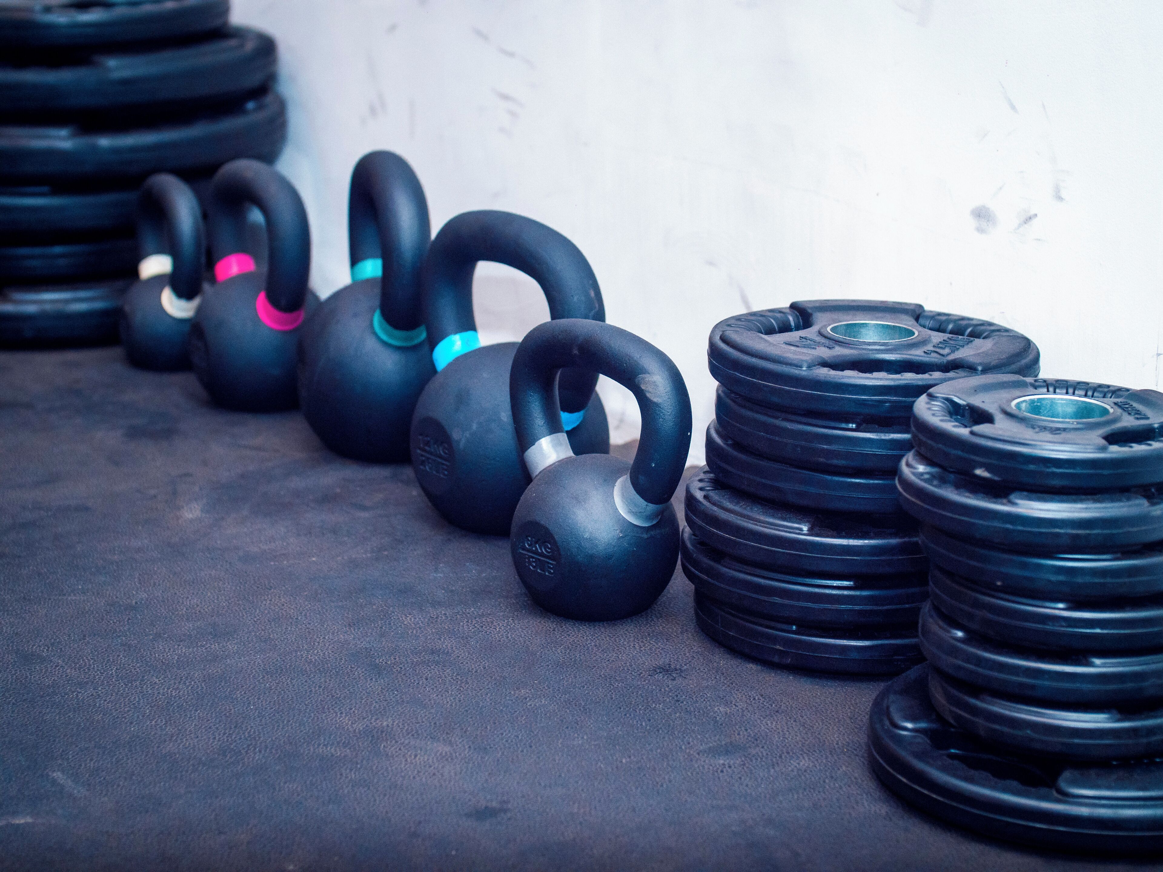 A row of black kettlebells with colored accents and a stack of weight plates on a concrete floor.
