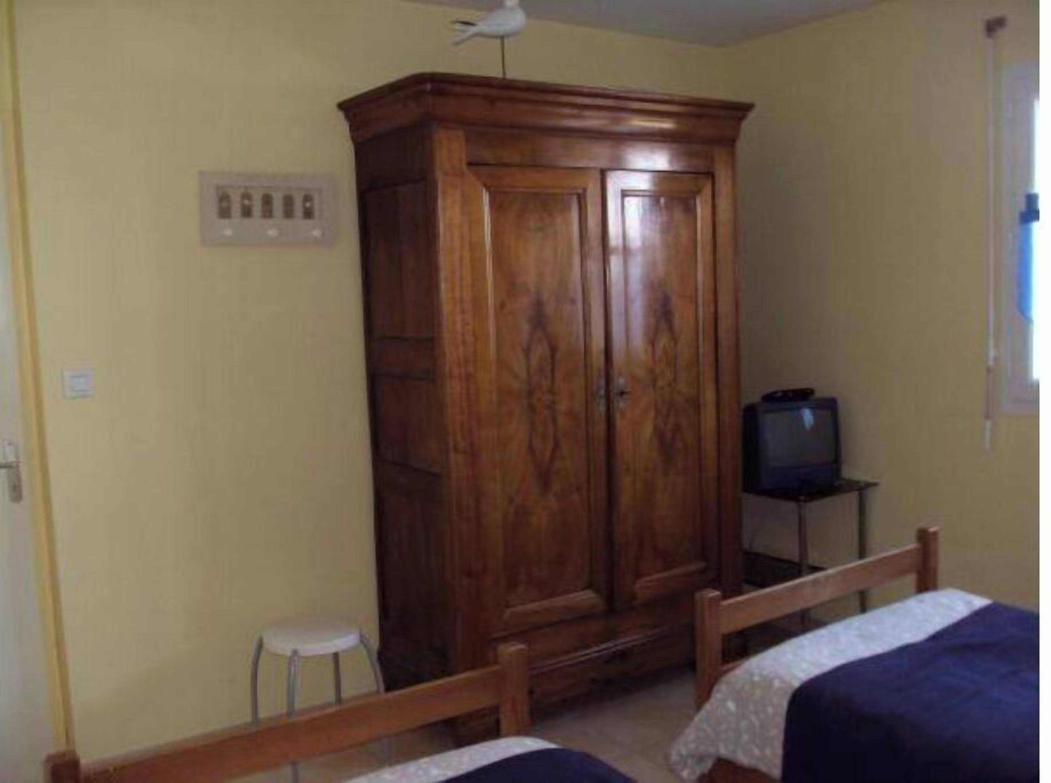 A room with a wooden wardrobe, a small stool, a bed with a blue blanket, and a television on a stand.