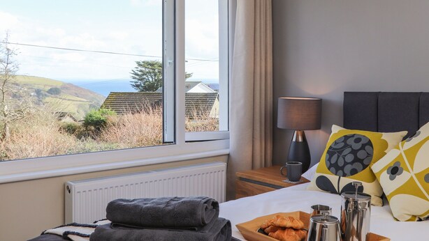 Ett hotellrum med ett stort fönster, en säng med kuddar, ett sängbord med en lampa och ett bricka med croissanter och en termos.