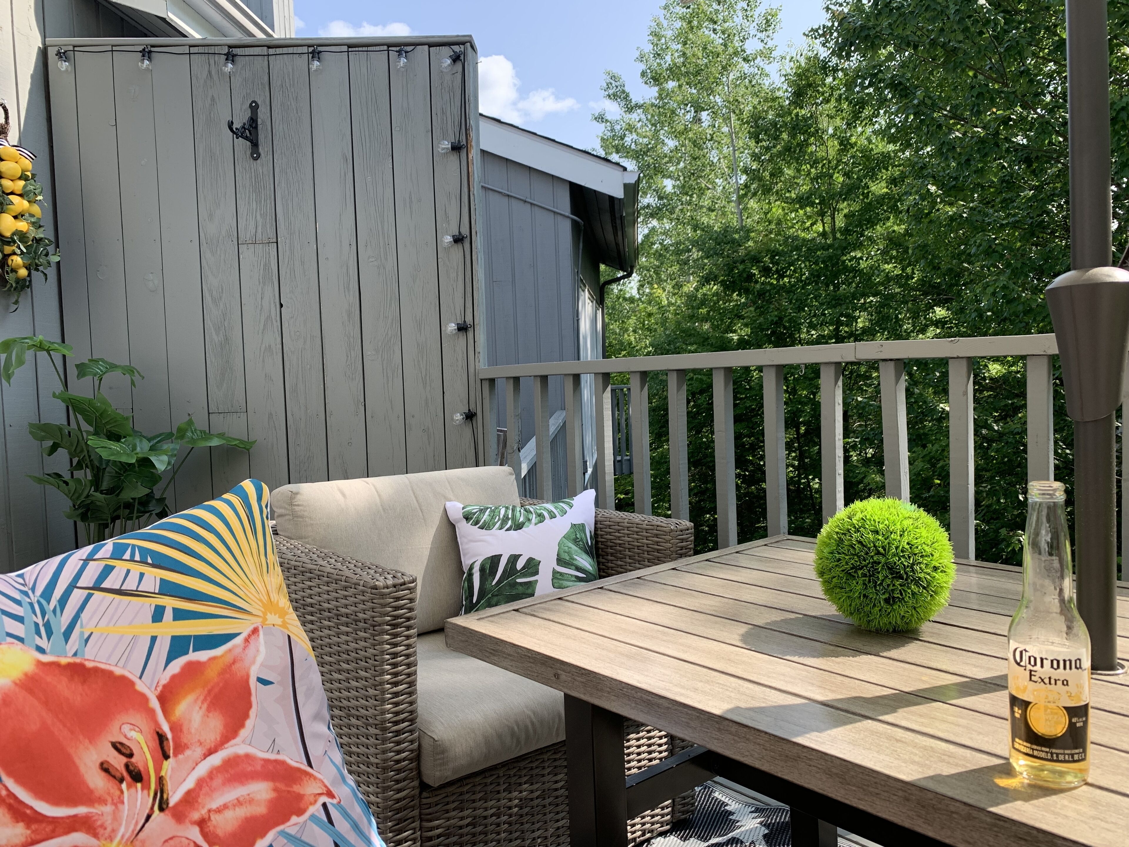 Eine Holzterrasse mit einer Korbus-Couch, einem runden Tisch und einer Flasche Corona Extra Bier.