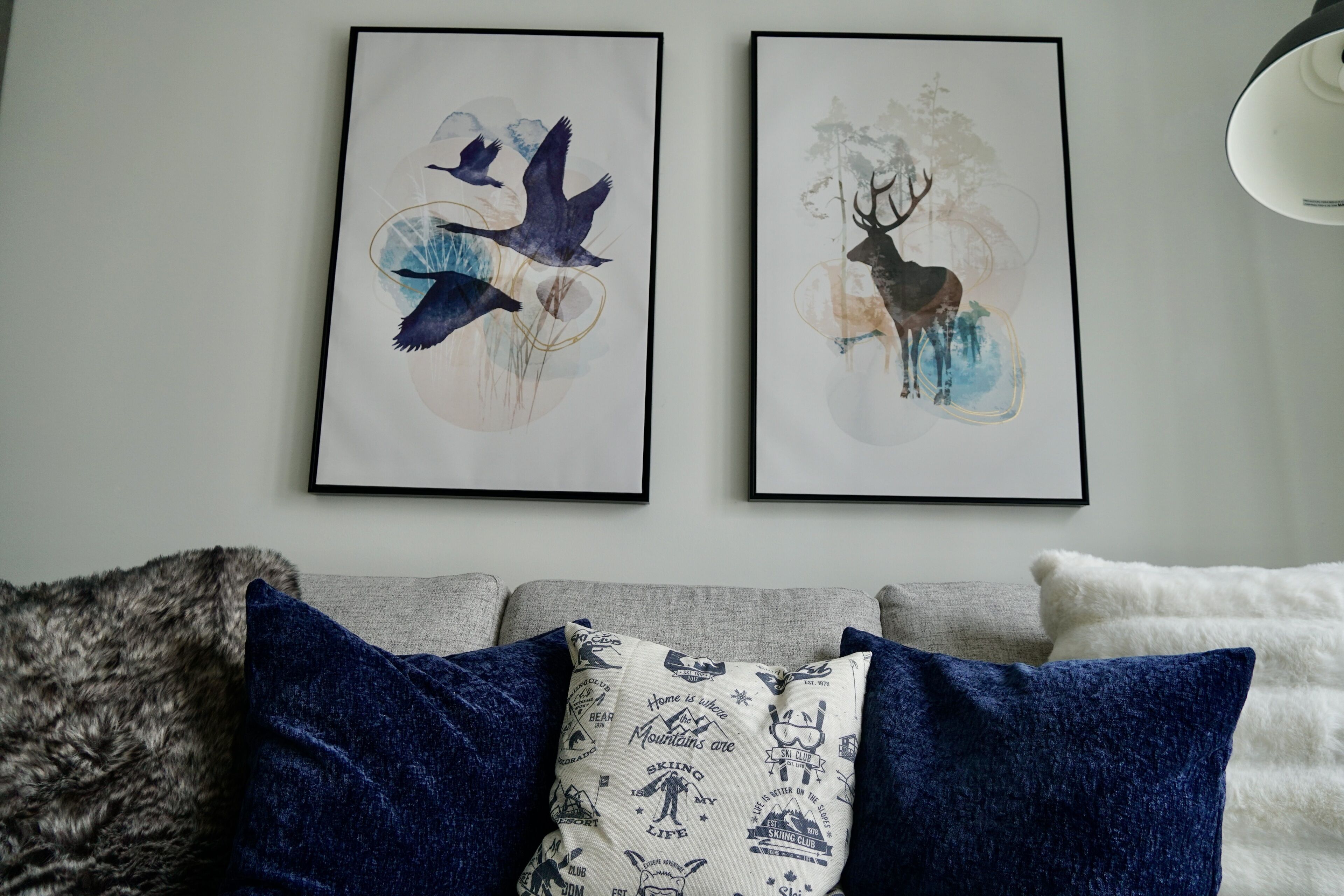 Two framed artworks depicting birds and a deer, a grey sofa with blue and white cushions, and a white throw blanket.