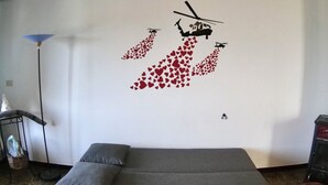 Una habitación con un sofá gris, un adhesivo mural con helicópteros y corazones, una lámpara de pie y una pequeña mesa con una cesta.