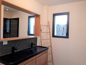 Une salle de bain avec un comptoir noir, un pare-feu en carrelage, un meuble-lavabo en bois et un grand miroir. Il y a une fenêtre donnant sur une clôture et une échelle appuyée contre le mur.
