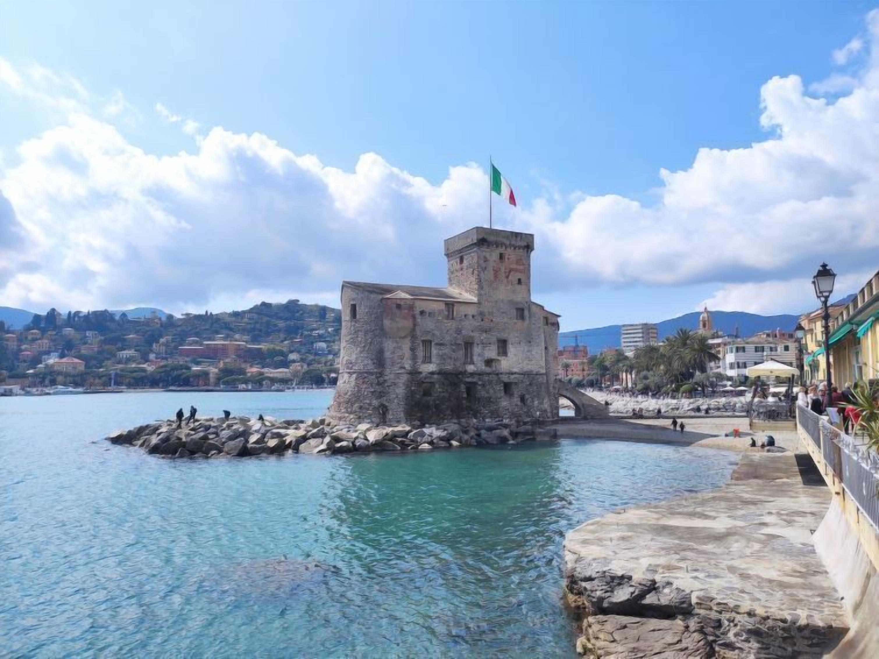Ein historischer Stein-Turm mit einer Flagge, eine Steinbrücke und eine Uferpromenade mit Gebäuden und blauem Himmel.