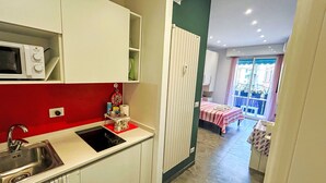 Uma cozinha compacta com micro-ondas, pia e uma parede vermelha de destaque. Uma varanda com mesa e cadeiras é visível através de uma porta aberta.