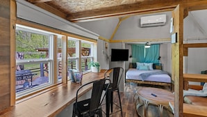 Ein Zimmer mit Holzboden, einem Bett mit blauer Vorhang, einem an der Wand montierten Fernseher und einem Fenster mit Blick auf einen Grillbereich.