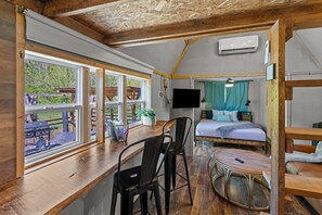 Una habitación con techo de madera, cama con cortina azul, televisor montado en la pared y ventana con vistas a una zona de barbacoa.