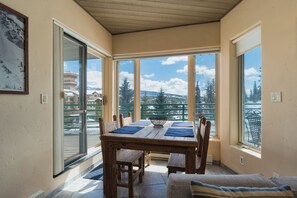 A dining area with a wooden table and chairs, large windows with a view of snow-covered mountains and trees, and a framed picture on the wall.