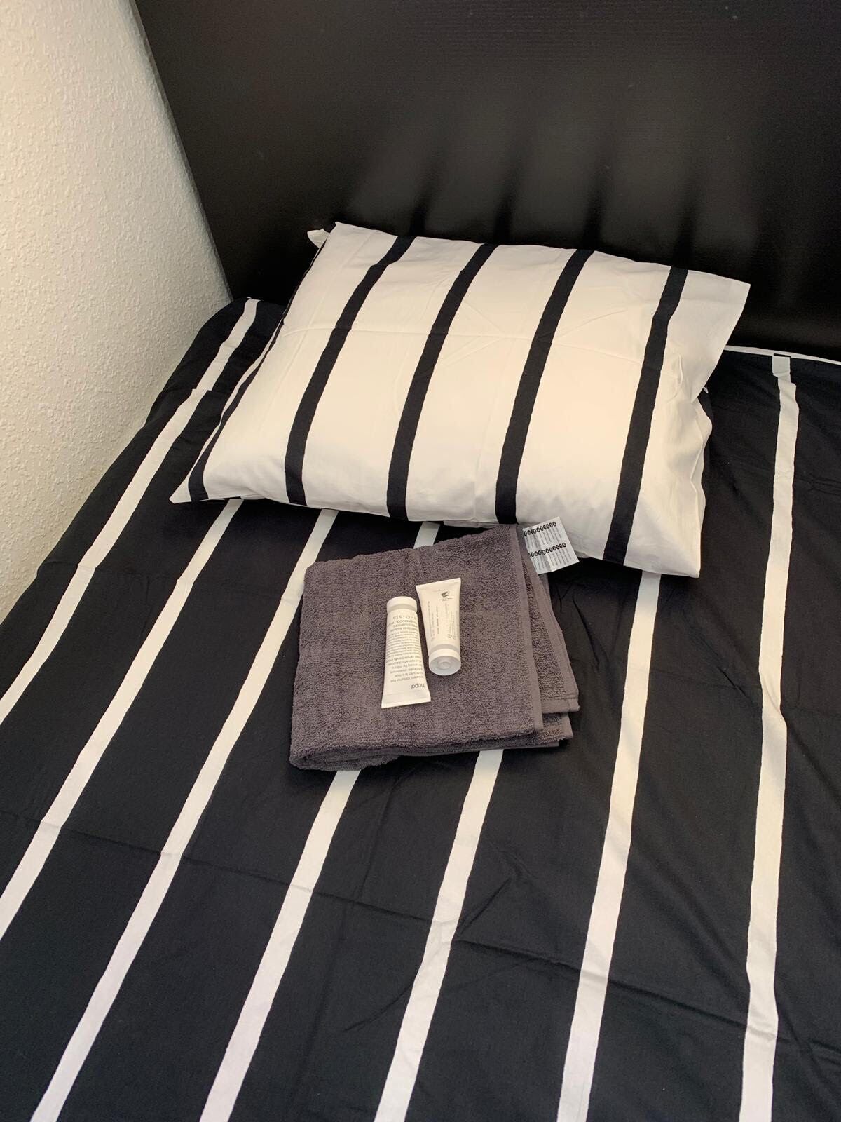 Un lit avec une literie à rayures noires et blanches, un oreiller à rayures et une serviette pliée, accompagnée d’un tube de crème et d’un petit paquet.