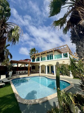 Un espace piscine avec un ciel d’un bleu éclatant, un bâtiment à deux étages doté d’un patio couvert et une végétation luxuriante tout autour.