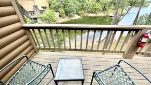 A wooden deck with two metal chairs and a small table, overlooking a lake with trees and a building in the background. - 80BPD, Four bedroom, two 1/2 bath log-sided condo with lake view , garage by RedAwning (Mount Ida)