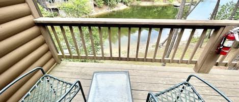 A wooden deck with two metal chairs and a small table, overlooking a lake with trees and a building in the background.