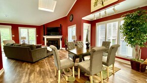 A living room with a fireplace, a dining area with chairs, and a large window with blinds. - 80BPD, Four bedroom, two 1/2 bath log-sided condo with lake view , garage by RedAwning (Mount Ida)