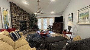 A cozy living room with a stone fireplace, a ceiling fan, and a television on a cabinet. - Forest view, log-sided two bedroom, two bath condo on Lake Ouachita. by RedAwning (Mount Ida)